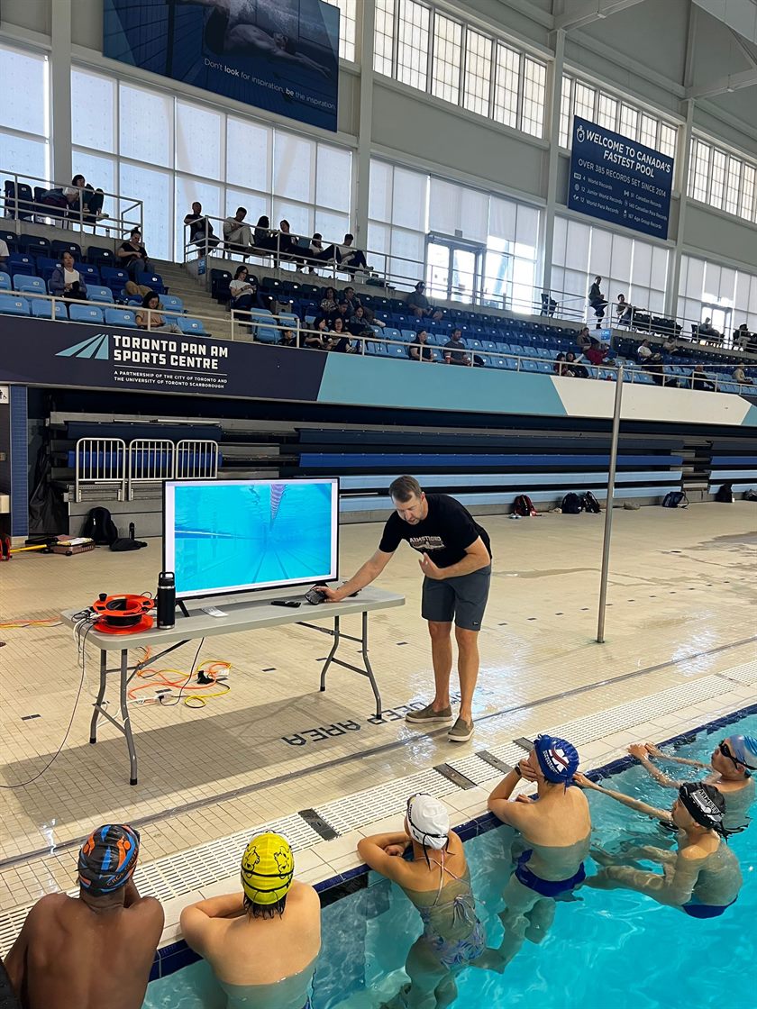 A coach reviewing underwater footage on a deck monitor while swimmers watch from the pool wall.