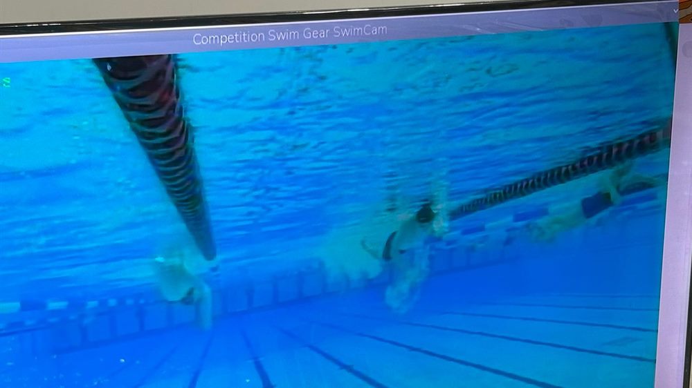 A deck monitor showing head-on underwater footage of swimmers coming down the lane.