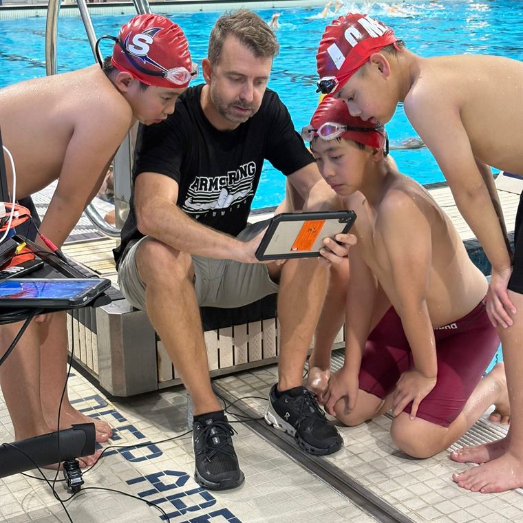 A coach on the pool deck showing tablet footage to three swimmers.