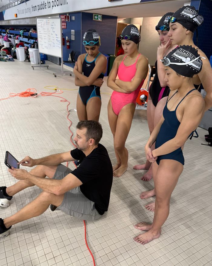 Young swimmers on the deck watching a replay on their coach's tablet.